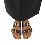 A close-up of a person's feet wearing black sandals with shiny brown and black crocodile-embossed straps and gold-tone buckles, styled with black trousers against a light gray background.