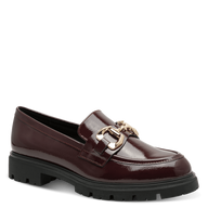 A single burgundy patent leather loafer with a chunky black lug sole and a gold-tone horsebit detail on the strap, shown against a solid green background.