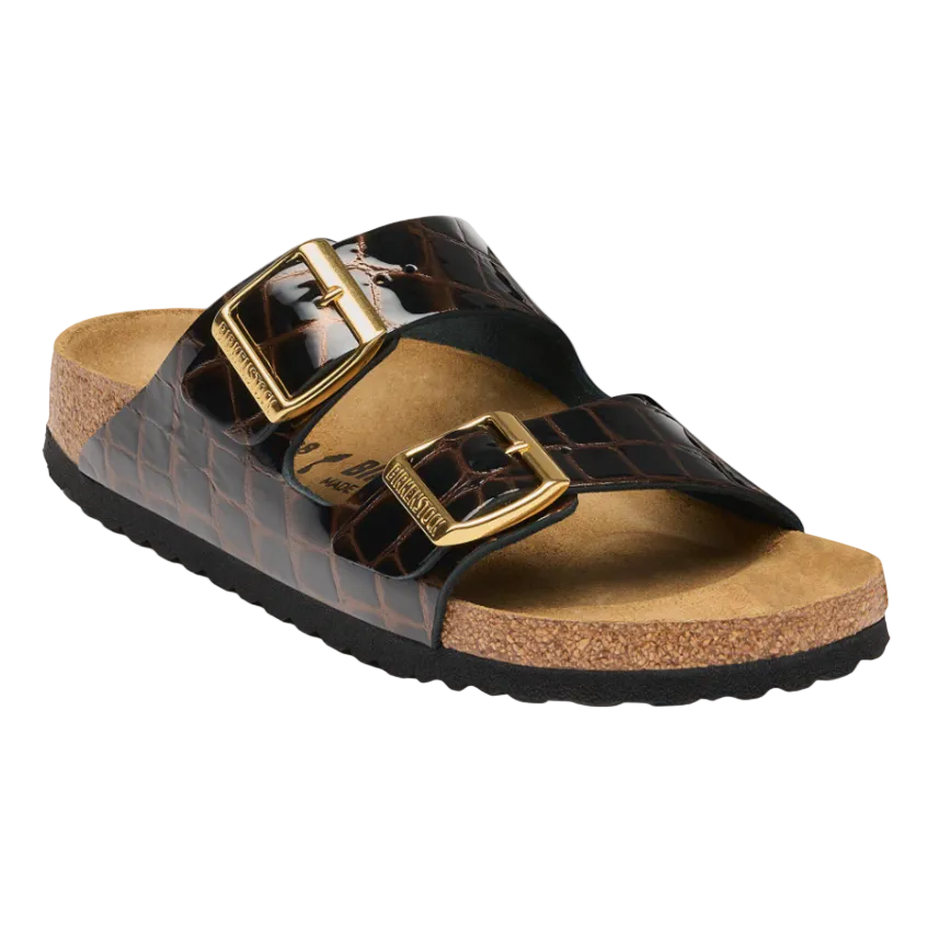 A pair of Birkenstock Arizona slide sandals in brown crocodile-embossed leather with gold-tone buckles, a tan cork footbed, and a black outsole, shown on a white background.